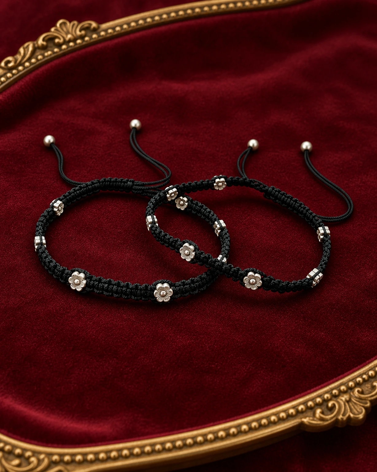 Pair of adjustable black thread anklets with silver flower charms on burgundy velvet.