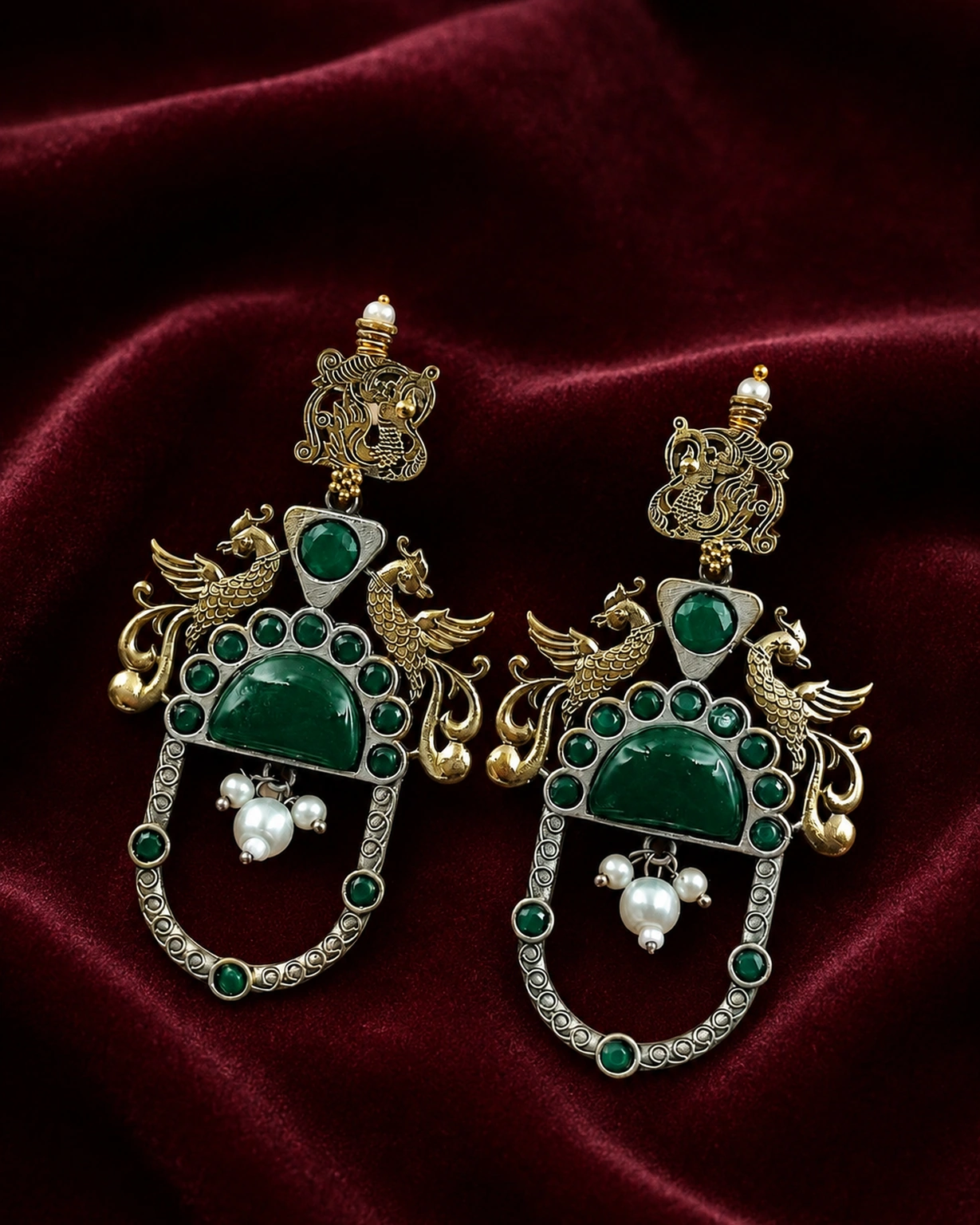 Green two-tone peacock earrings displayed on rich burgundy velvet.